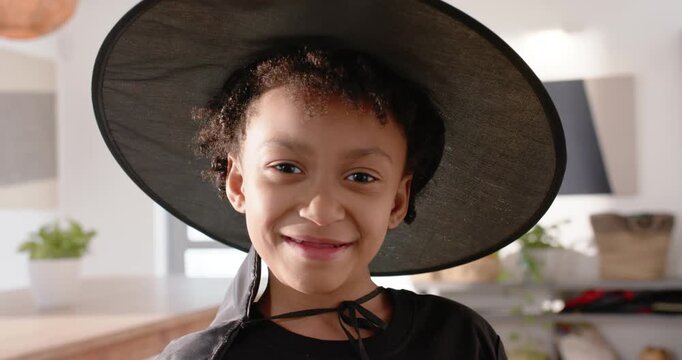 Camera pulling back African American child in kitchen in witch hat showing pumpkin pail for candy