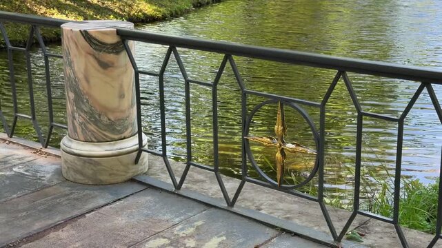 Wrought iron bridge railing with geometric patterns and a solid stone pillar alongside a park river. Bright sunlight casting shadows on a peaceful summer day.