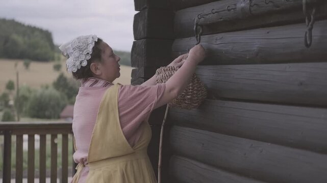 woman in traditional peasant costume, yellow pinafore dress, pink blouse, patterned scarf, lace headpiece, Historical portrait, holding wooden stick, near dark log cabin wall, basket, rural heritage