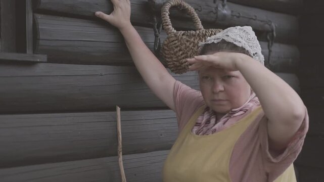 woman in traditional peasant costume, yellow pinafore dress, pink blouse, patterned scarf, lace headpiece, Historical portrait, holding wooden stick, near dark log cabin wall, basket, rural heritage