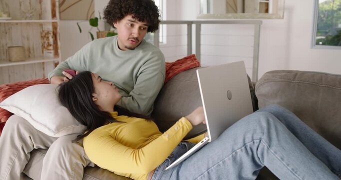 Diverse couple reclining on couch woman typing on laptop as man leaning from phone watching screen