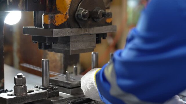 Industrial worker operating metal stamping press factory workshop, wearing blue protective clothing and white glove while positioning sheet material during metal manufacturing process