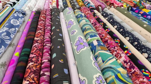 parallel rows of rolled fabric bolts on retail rack, featuring floral, geometric, and abstract printed patterns in vibrant purple, pink, teal, green, navy, and bolt