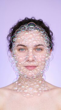 Young woman with a face mask made of iridescent soap bubbles on a purple background creates a playful surrealist portrait with shimmering soap suds covering her skin and neck.