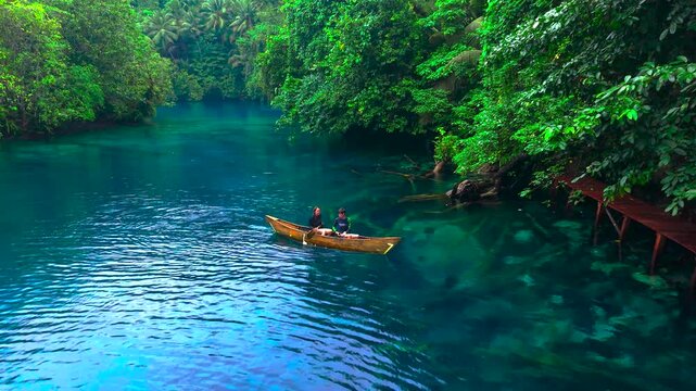 Aerial view of a young woman and a young man paddling a wooden canoe on clear blue water near tropical forest at Paisupok Lake, Indonesia.