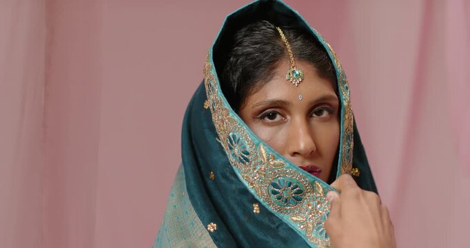 Indian woman camera cueing, lifting teal shawl with right hand, pink backdrop revealing maang tikka
