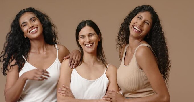 Diverse female friends posing closer, touching center woman's arms, laughing in studio tan backdrop