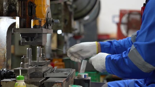 Industrial worker operating metal stamping machine factory workshop, wearing protective blue glove and clothing while shaping precision parts with focused manufacturing skill indoors during