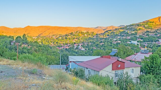 A high angle panoramic view of Bitlis city center, Turkey. The image features a bare mountain illuminated by golden sunlight at sunset or sunrise, with local houses nestled among lush green trees in t