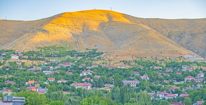 A high angle panoramic view of Bitlis city center, Turkey. The image features a bare mountain illuminated by golden sunlight at sunset or sunrise, with local houses nestled among lush green trees in t
