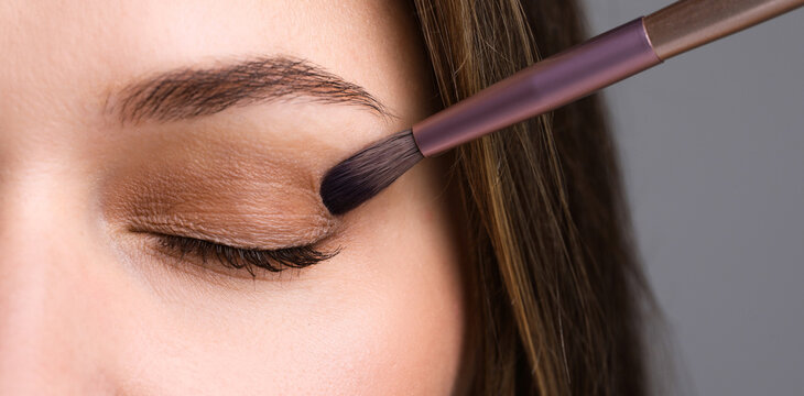 Applying nude eye shadow with makeup brush onto woman's face against light background, closeup