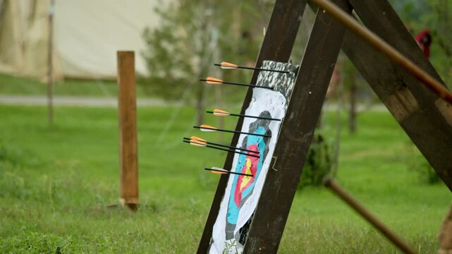 Outdoor archery range scene shows standard target mounted on wooden frame. Point view perspective highlights incoming arrow entering colored target zones near bullseye area.