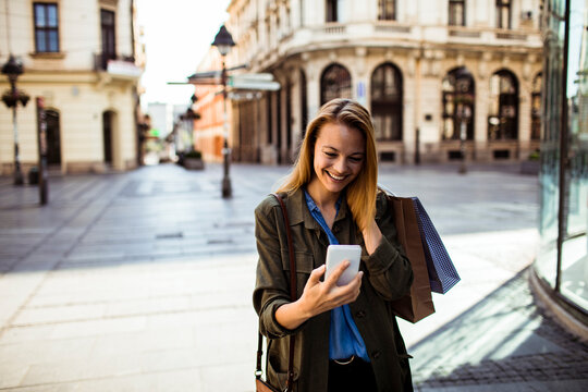 Smiling woman checking smartphone with shopping bags on historic city street