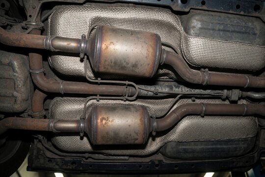 Underside view of car exhaust system components. Catalytic converters and pipes are visible on vehicle chassis, part of emission control assembly.