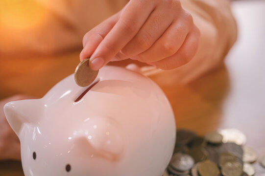 Young girl putting gold coin in a white ceramic piggy bank with both hands over wooden table in concept of saving money and financial investment for future and retirement.