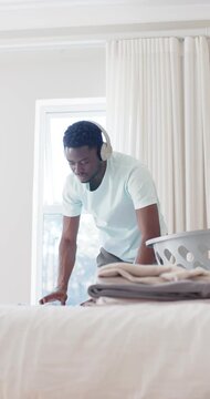 Vertical video: African American man entering bedroom, folding clothes from basket on bed to tidy