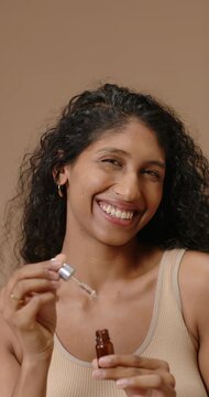 Vertical video: Indian woman opening amber dropper bottle placing serum on cheek for skin in studio