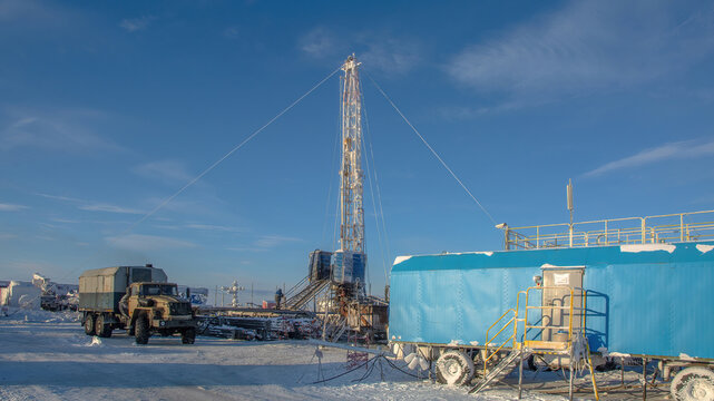 An oil and gas well workover rig operating in harsh Arctic winter conditions. Industrial landscape featuring heavy machinery, snow-covered permafrost, and a sunny clear polar day.