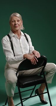 Vertical video: Senior woman resting chin, looking at camera, raising hand on black chair in studio