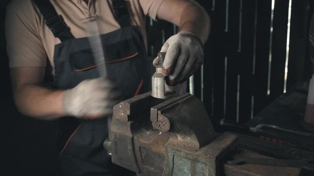 Skilled technician in work gloves operates a metal vise, securing a cylindrical object for precise adjustments in a dimly lit workshop with tools and equipment in the background