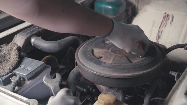 Auto mechanic in gray overalls prepares to replace air filter inside a car engine compartment, with tools and equipment visible in the garage background