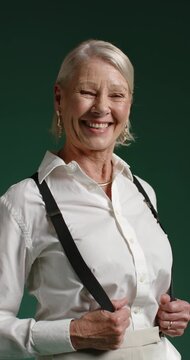 Vertical video: Senior woman holding suspenders, laughing, tilting head at studio for camera