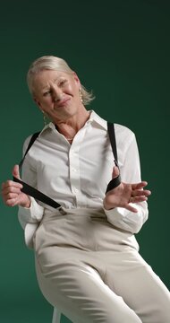 Vertical video: Senior female playing with suspenders, posing and smiling for camera on green set