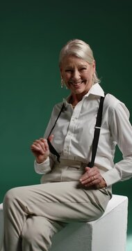 Vertical video: Camera rolling, senior female posing on white cube in studio, adjusting suspenders