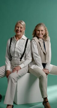 Vertical video: Two mature women sitting on white cube in studio, responding to prompt