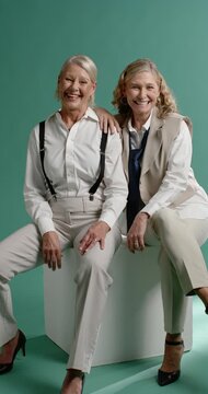 Vertical video: Two mature women posing in studio, smiling and leaning on white cube, wearing tie