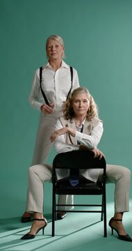 Vertical video: On cam senior women posing, sitting chair, standing adjusting suspenders for shoot