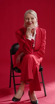 Vertical video: Senior woman in red-suit on black folding chair in studio, lifting chin, smiling