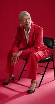 Vertical video: Senior woman in red suit sitting on black chair in studio after cue adjusting gaze