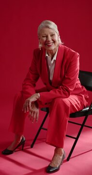 Vertical video: Senior woman noticing camera leaning forward posing in red suit on chair in studio