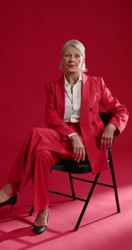 Vertical video: Senior woman sitting chair looking as camera panning posing in red suit on red set