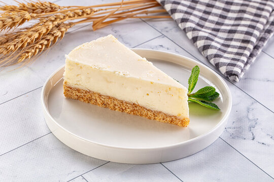 Slice of creamy cheesecake with graham cracker crust on white plate, garnished with fresh mint, set against marble surface with wheat stalks and checkered napkin