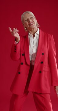 Vertical video: Senior woman smiling at camera fixing red suit and dancing confidently in studio