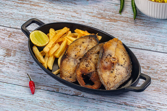 Grilled kingfish steak with golden fries and lemon wedge served in black oval dish on rustic wooden table with red and green chili peppers