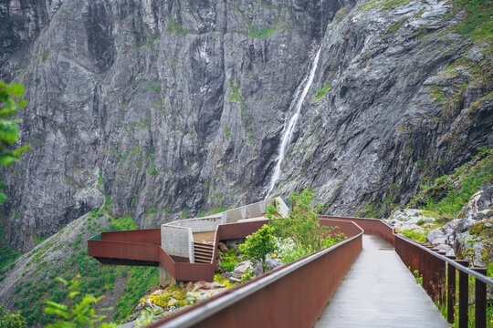 Modern steel viewing platform at Trollstigen Norway featuring a mountain waterfall backdrop and rocky terrain.