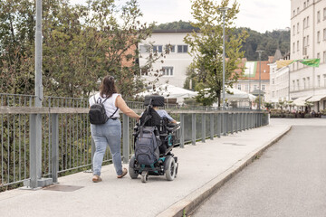 Obraz premium Powered wheelchair user moving along an outdoor walkway accompanied by a Personal assistant, supporting independent mobility.