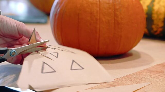 Hands use scissors to cut triangular shapes from a paper pumpkin face template beside an orange pumpkin, Halloween craft, seasonal decoration.