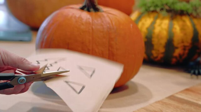 Hands cut a paper face template with scissors beside an orange pumpkin, preparing a stencil for decoration, Halloween craft, pumpkin decorating.