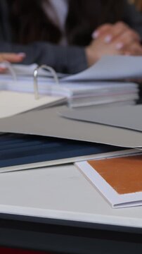 Hands sorting binder pages on office table, flipping documents and reviewing paper samples with ring binders, stacked folders, notebooks, pens, and loose sheets during collaborative work.