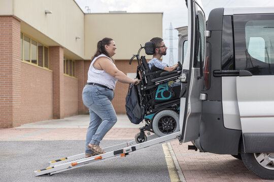 Personal assistant assisting a powered wheelchair user down a vehicle ramp for safe and accessible transport, side view.