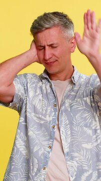Middle-aged man covering ears with hands and shaking head in clear rejection, refusing to hear or accept something unpleasant. Young guy expressing protest and discomfort isolated on yellow background