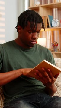 African American young man on home sofa reading open book with curious focus. Black boy on home sofa turning paper page enjoying calm study rest education hobby in warm cozy home living room interior.