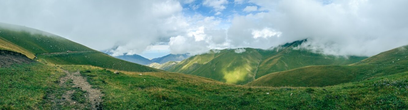 Superwide Panorama view of rolling green hills under cloudy sky. Mist blankets valleys, softening distant mountain ridges. Lush vegetation covers slopes. Kyrgyzstan Valley of Tranquility Trail Hiking