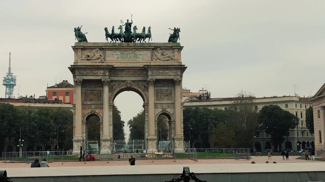 A scenic panning shot of the historic Arco della Pace triumphal arch with classical bronze statues and people relaxing in Piazza Sempione on a cloudy day in Milan.

