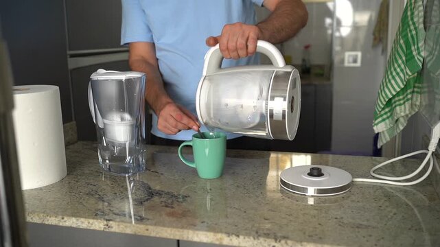 Man making a cup of tea in the morning, pouring boiling water from an electric kettle into a mug
