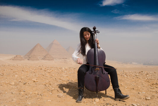Focused young woman cellist actively bowing a cello while seated on the sandy Giza plateau with the iconic Great Pyramids of Giza dissolving into